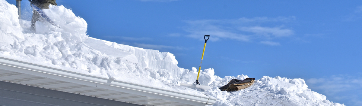 Un toit québécois recouvert d’une épaisse couche de neige, avec une pelle jaune enfoncée dans la neige sous un ciel bleu clair. Cette image illustre comment le climat québécois peut affecter votre toiture, nécessitant un entretien hivernal régulier.